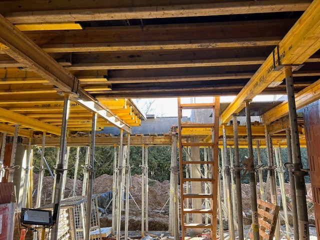 Wnętrze budynku w trakcie budowy, widoczne żółte szalunki stropowe wsparte na metalowych stemplach, drabina oparta o konstrukcję, w tle drzewa i ziemia.