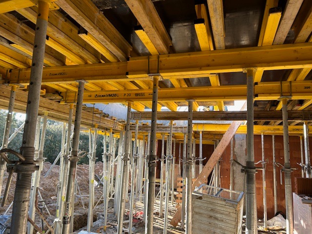 Szare metalowe podpory podtrzymujące żółte drewniane szalunki stropowe w trakcie budowy domu z cegły.