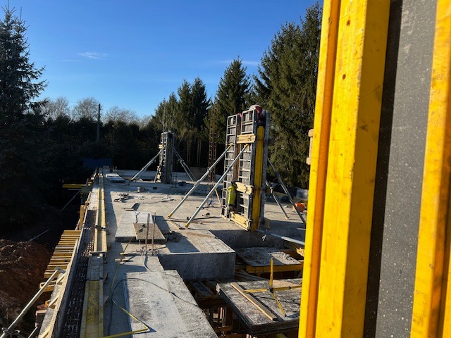 Wylany fundament pod budynek z drewnianym szalunkiem i stalowym zbrojeniem, widoczne żółte elementy konstrukcyjne i pracownik w oddali.