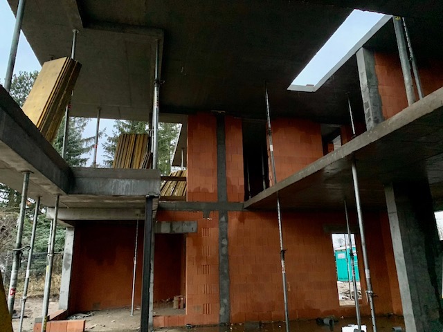 Surowy stan budynku w trakcie budowy, widoczne czerwone cegły, betonowe stropy i podpory oraz deski szalunkowe, zarys okien i otworów drzwiowych, na zewnątrz drzewa.