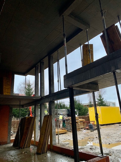 Wnętrze surowego budynku w trakcie budowy z żelbetową konstrukcją stropu, widoczne podpory i szalunki, na zewnątrz plac budowy z kontenerem i ekipą budowlaną.