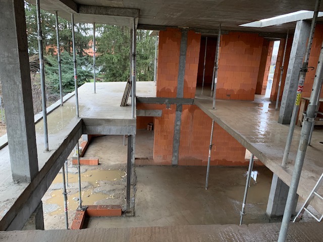 Surowy stan budowy dwukondygnacyjnego domu murowanego z widocznymi podporami stalowymi, mokrą podłogą i częściowo wzniesionymi ścianami z czerwonej cegły.