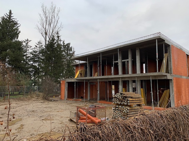 Dwukondygnacyjny budynek w stanie surowym otwartym z czerwonej cegły, widoczne szalunki i podpory, teren budowy z błotem i materiałami budowlanymi, drzewa w tle.