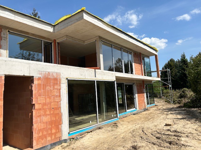 Budowa dwukondygnacyjnego domu z czerwonej cegły i dużymi oknami w Warszawie, widok z zewnątrz na plac budowy i rusztowanie.