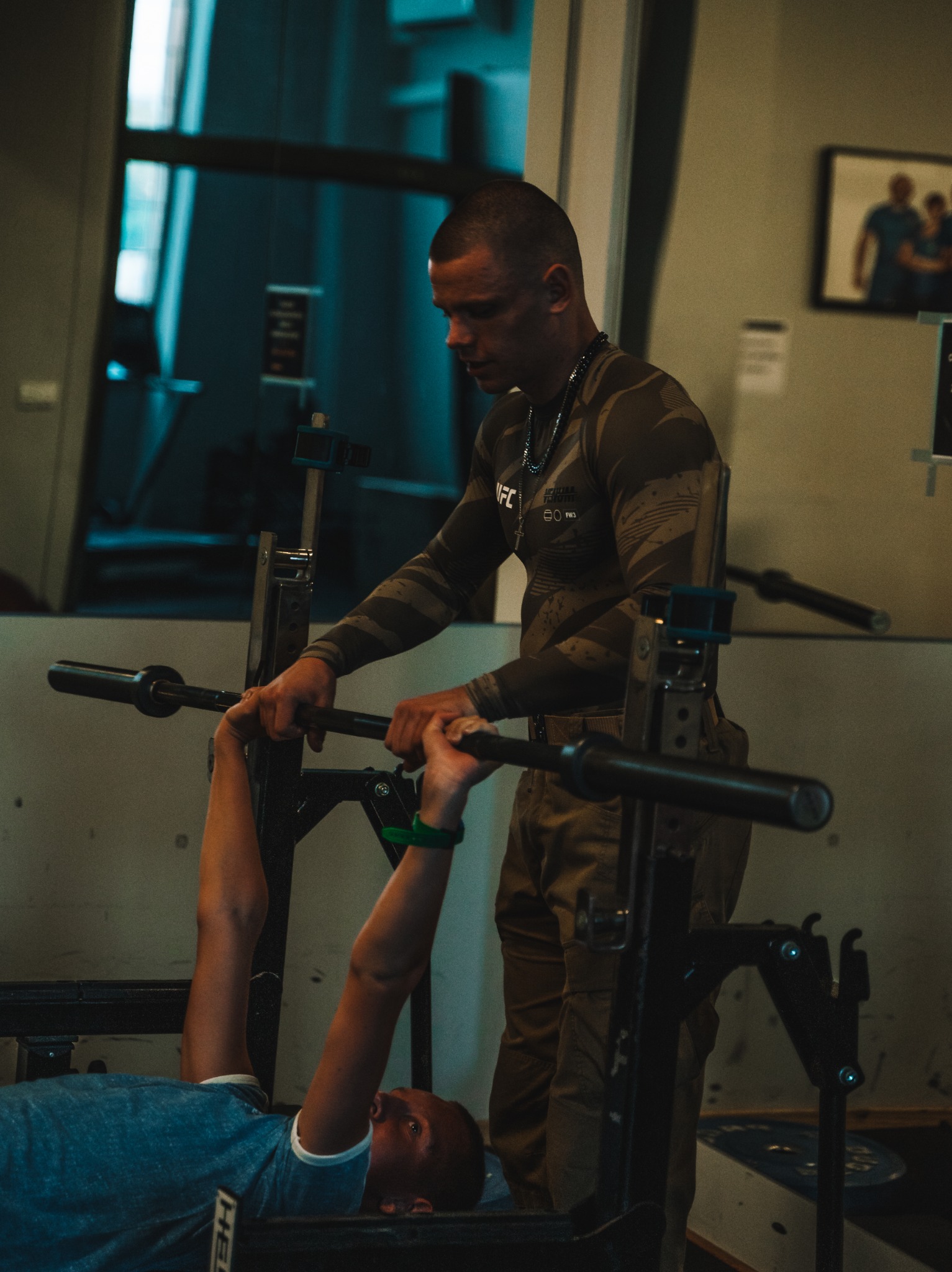 Mężczyzna w koszulce moro asekuruje ćwiczącego mężczyznę leżącego na ławeczce podczas wyciskania sztangi w siłowni.