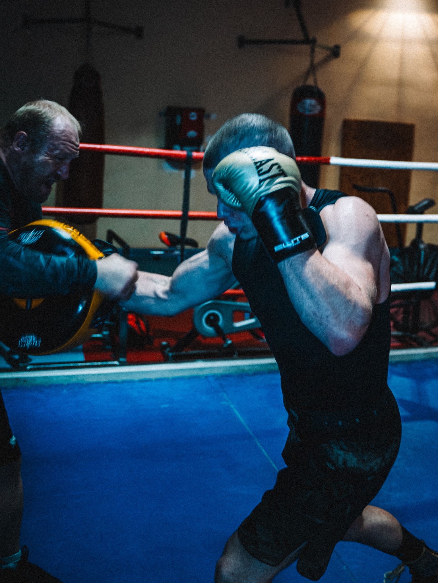 Zawodnik wyprowadza cios w rękawicach bokserskich na tle ringu, trener trzyma tarczę treningową
