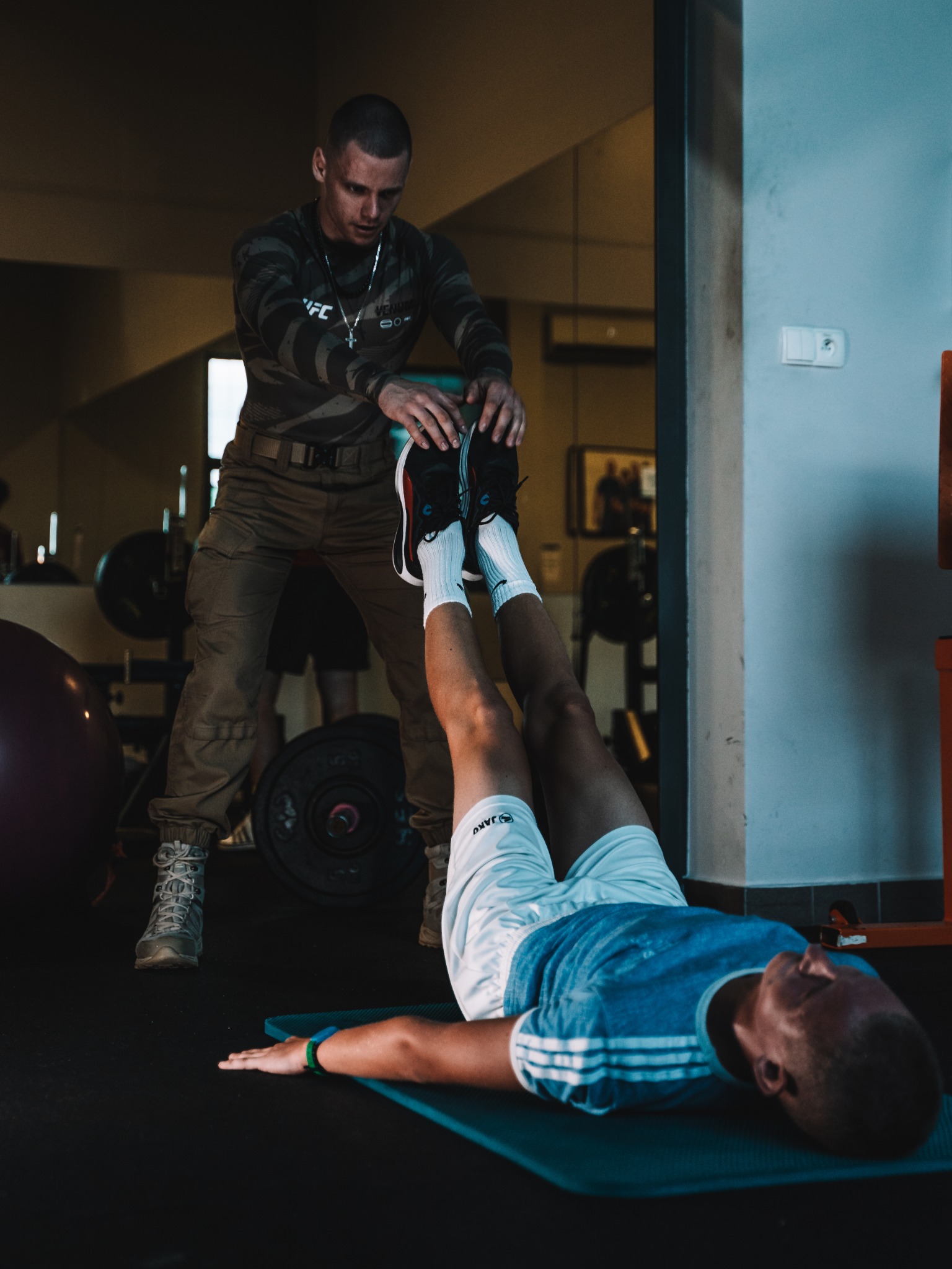 Mężczyzna w stroju moro pomaga drugiemu mężczyźnie leżącemu na macie podnosić nogi w siłowni, widoczne obciążenia i sprzęt do ćwiczeń.