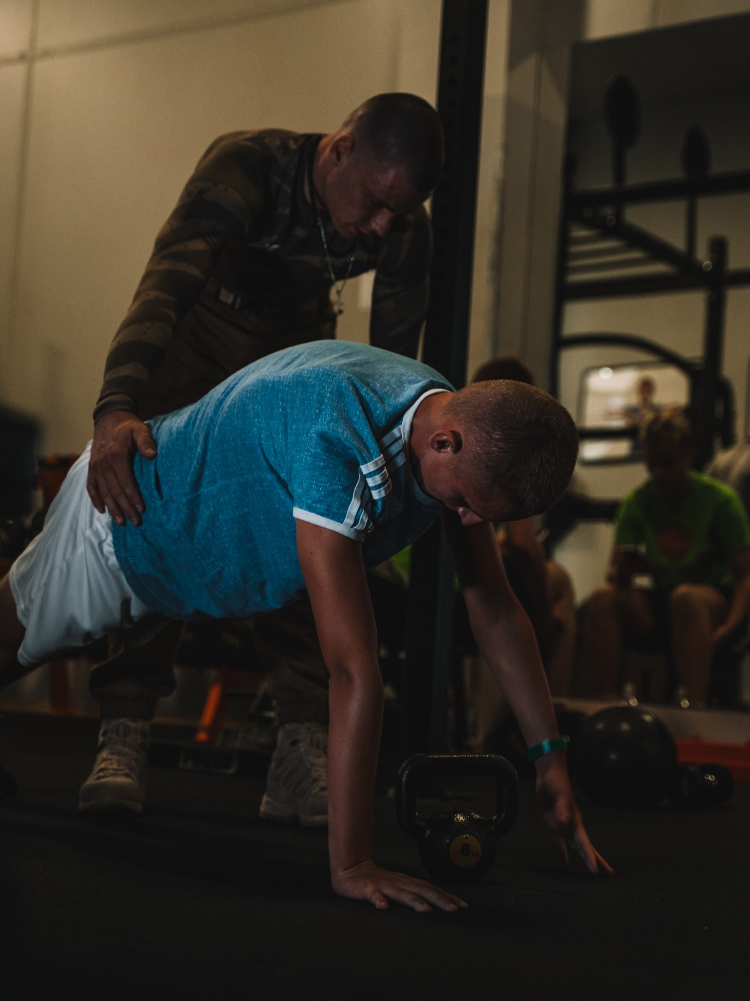 Mężczyzna w kamuflażowej koszuli koryguje pozycję drugiego mężczyzny podczas robienia pompek w siłowni, z kettlebellem widocznym na podłodze.