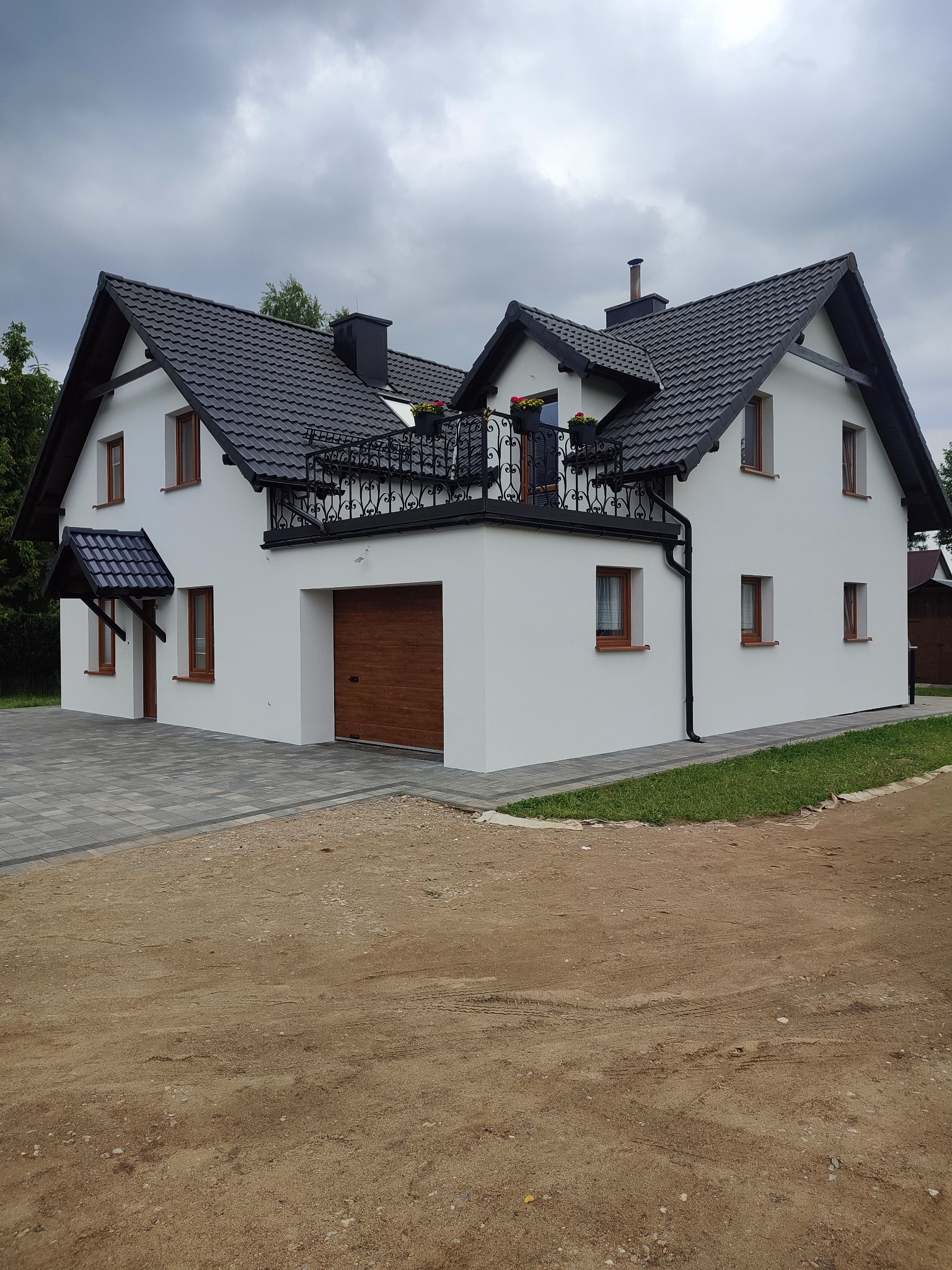 Biały dom z ciemną dachówką i balkonem z kutą balustradą, widok z zewnątrz. Nowoczesna architektura z drewnianymi oknami i garażem. Brukowany podjazd i fragment trawnika.
