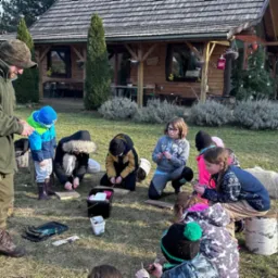 Warsztaty dla dzieci na trawie przed drewnianym domkiem. Animator w zielonej kurtce instruuje grupę dzieci. Obok leży pies. W tle las i choinki.
