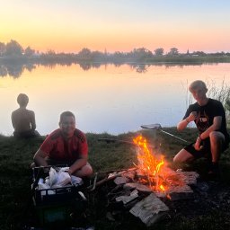 LASOMIR - Młodzi ludzie siedzą nad jeziorem przy ognisku o zachodzie słońca. Jeden z nich siedzi bez koszulki nad brzegiem, a pozostali obok ogniska.
