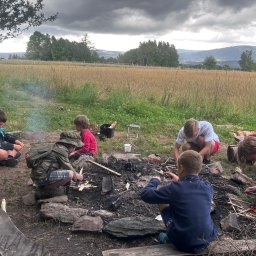 LASOMIR - Grupa dzieci rozpala ognisko na łące, w tle las i góry pod pochmurnym niebem. Ognisko otoczone kamieniami, dzieci siedzą wokół, używając noży i krzesiwa.