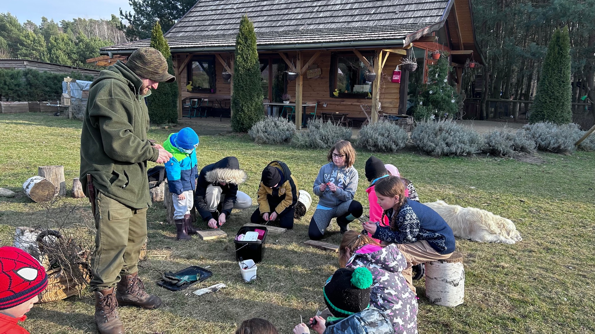 Warsztaty dla dzieci na trawie przed drewnianym domkiem. Animator w zielonej kurtce instruuje grupę dzieci. Obok leży pies. W tle las i choinki.