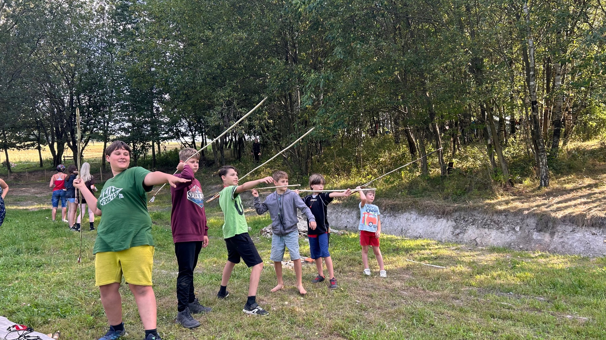 Grupa dzieci celuje patykami w leśnej scenerii podczas zabawy na świeżym powietrzu, prawdopodobnie w ramach zorganizowanej imprezy.