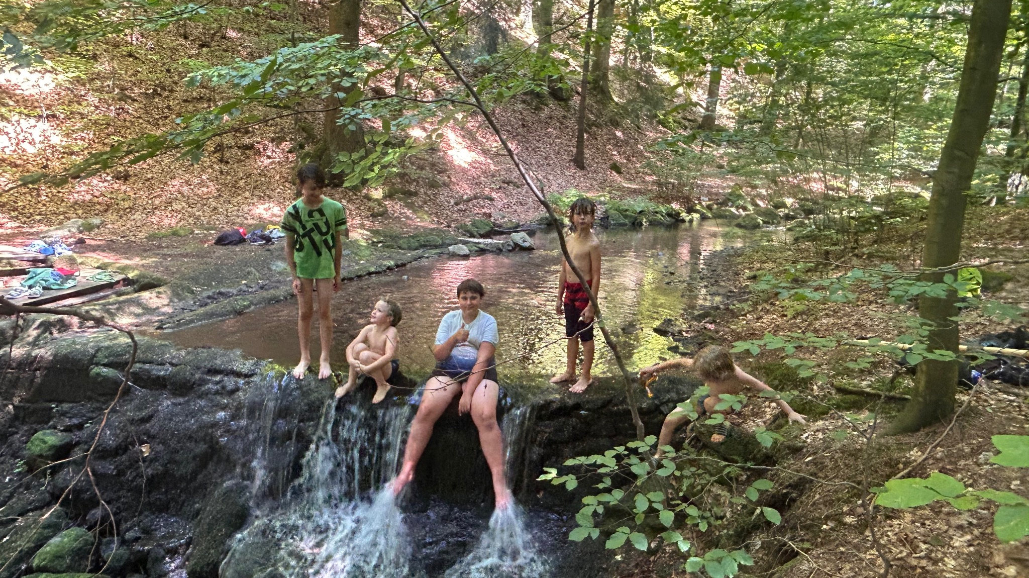 Grupa dzieci bawi się w leśnym strumieniu, siedząc i stojąc na skalnym wodospadzie. W tle widoczny las i ubrania. Jeden chłopiec wspina się po skałach.