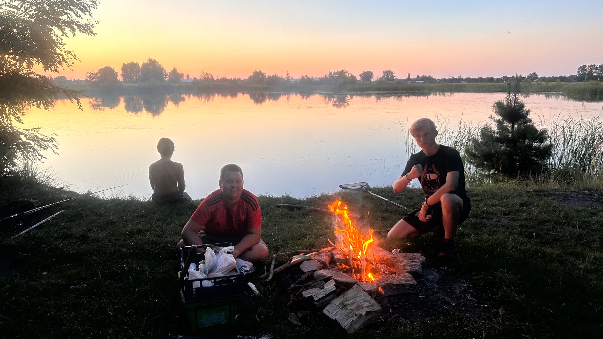 Młodzi ludzie siedzą nad jeziorem przy ognisku o zachodzie słońca. Jeden z nich siedzi bez koszulki nad brzegiem, a pozostali obok ogniska.