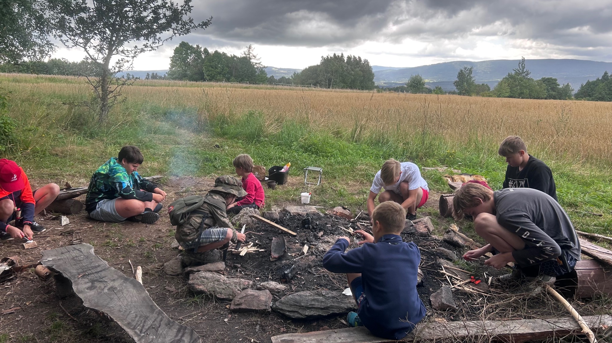 Grupa dzieci rozpala ognisko na łące, w tle las i góry pod pochmurnym niebem. Ognisko otoczone kamieniami, dzieci siedzą wokół, używając noży i krzesiwa.