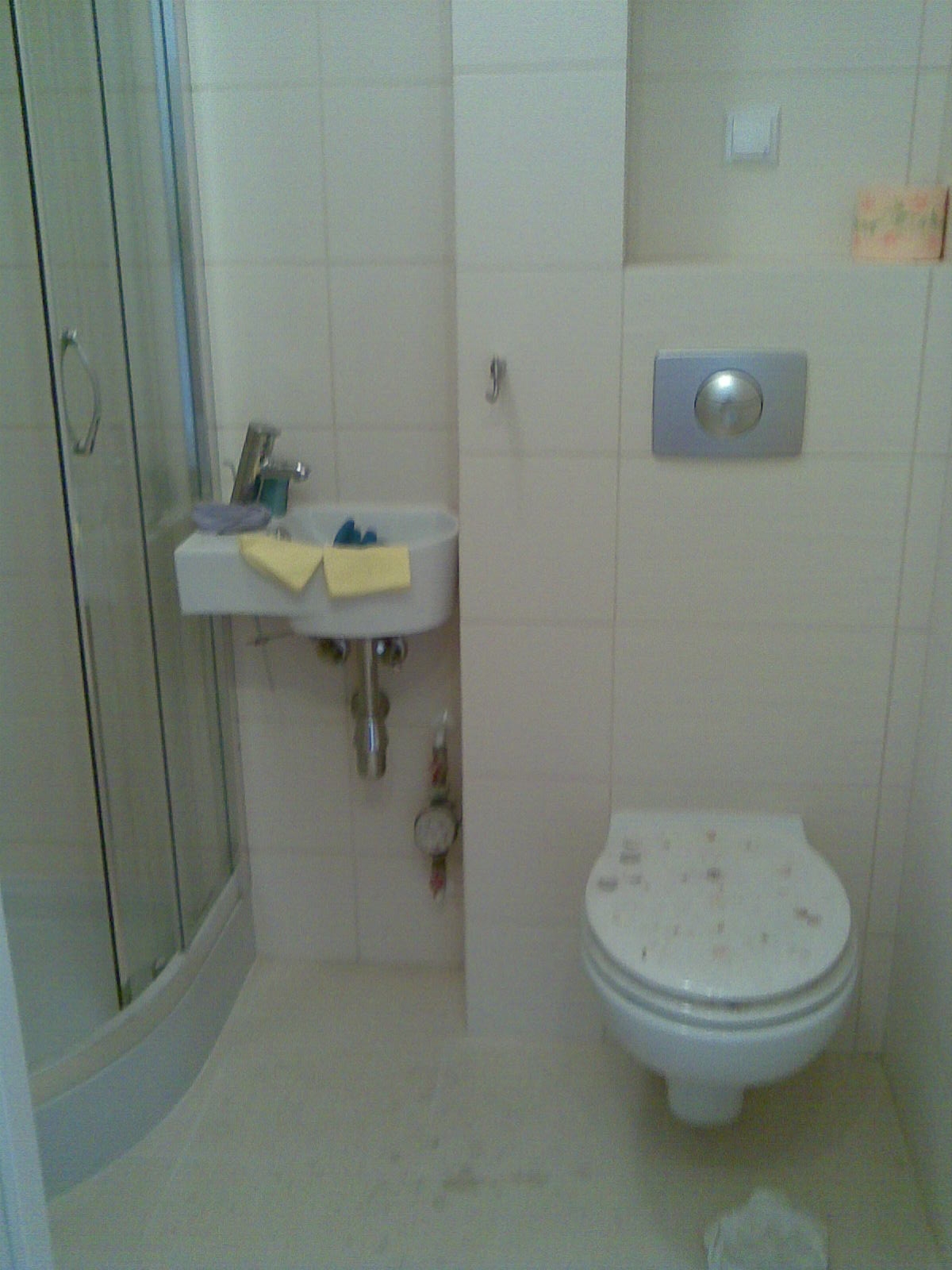 Niewielka łazienka z kabiną prysznicową, umywalką naścienną z baterią, toaletą podwieszaną i białymi płytkami. Na umywalce leżą żółte ściereczki. Na podłodze widoczne plamy.
