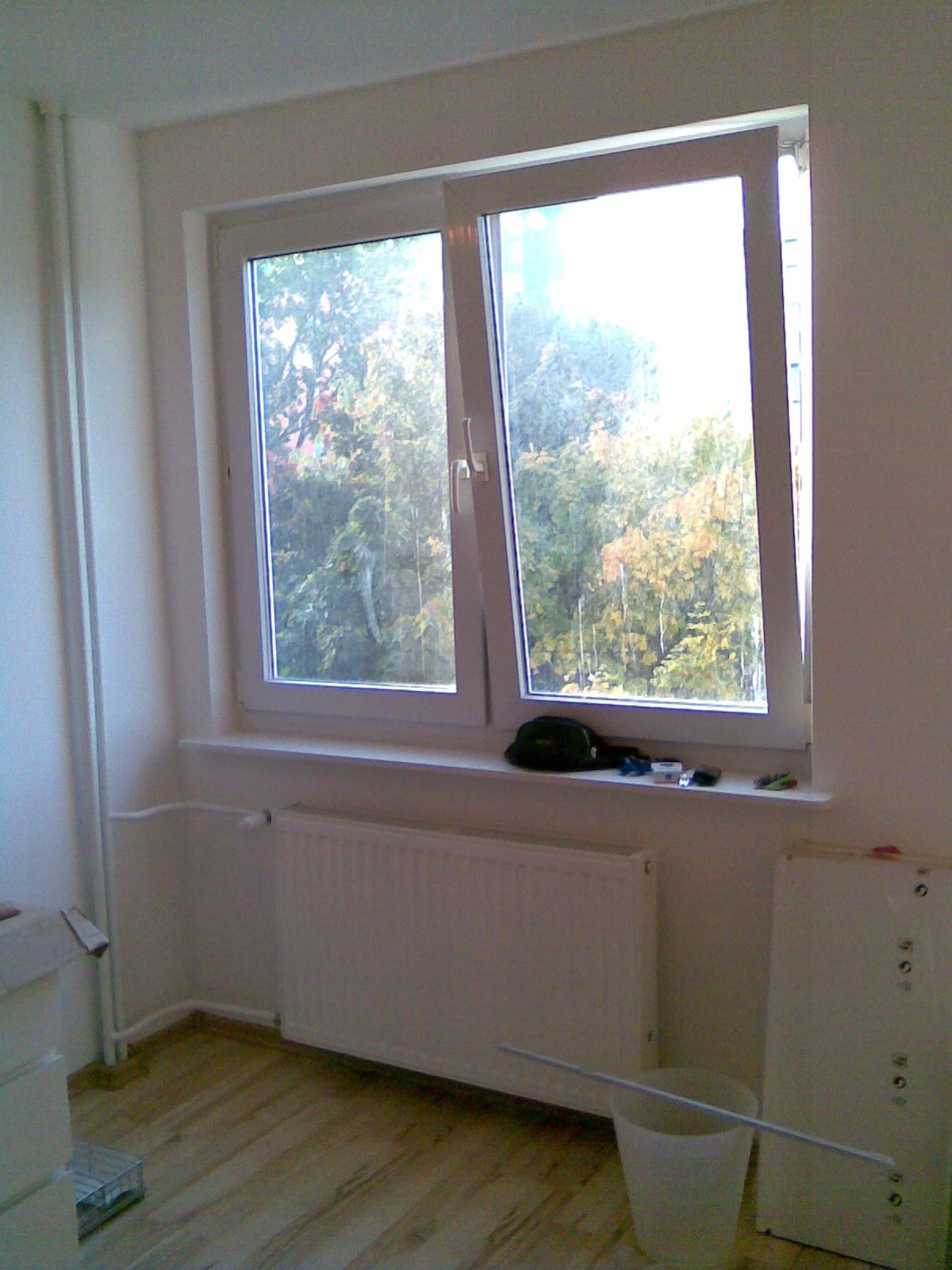 Pokój po remoncie, widoczne nowe okno PCV, grzejnik oraz drewniana podłoga. Na parapecie leżą drobne przedmioty.