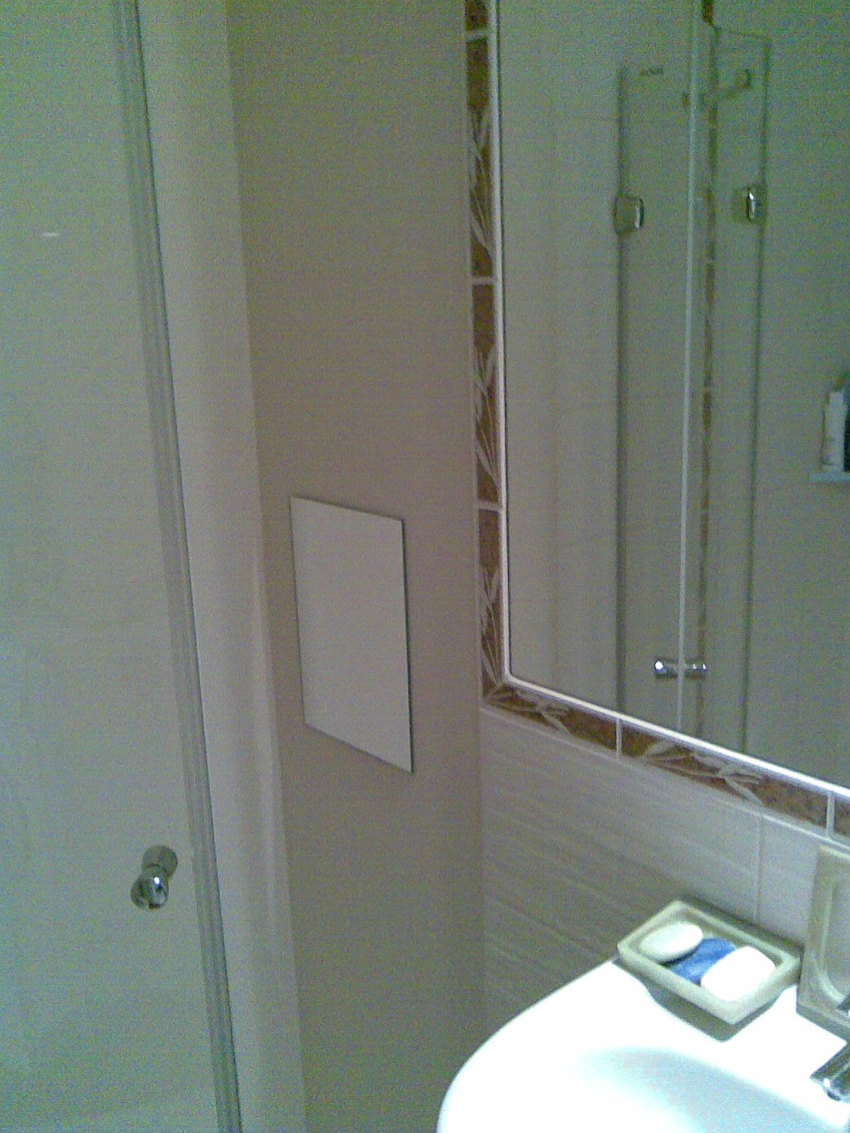 Wykończona łazienka z białymi płytkami, lustrem z dekoracyjną ramą i ukrytym dostępem do instalacji za pomocą białych drzwiczek rewizyjnych na ścianie.