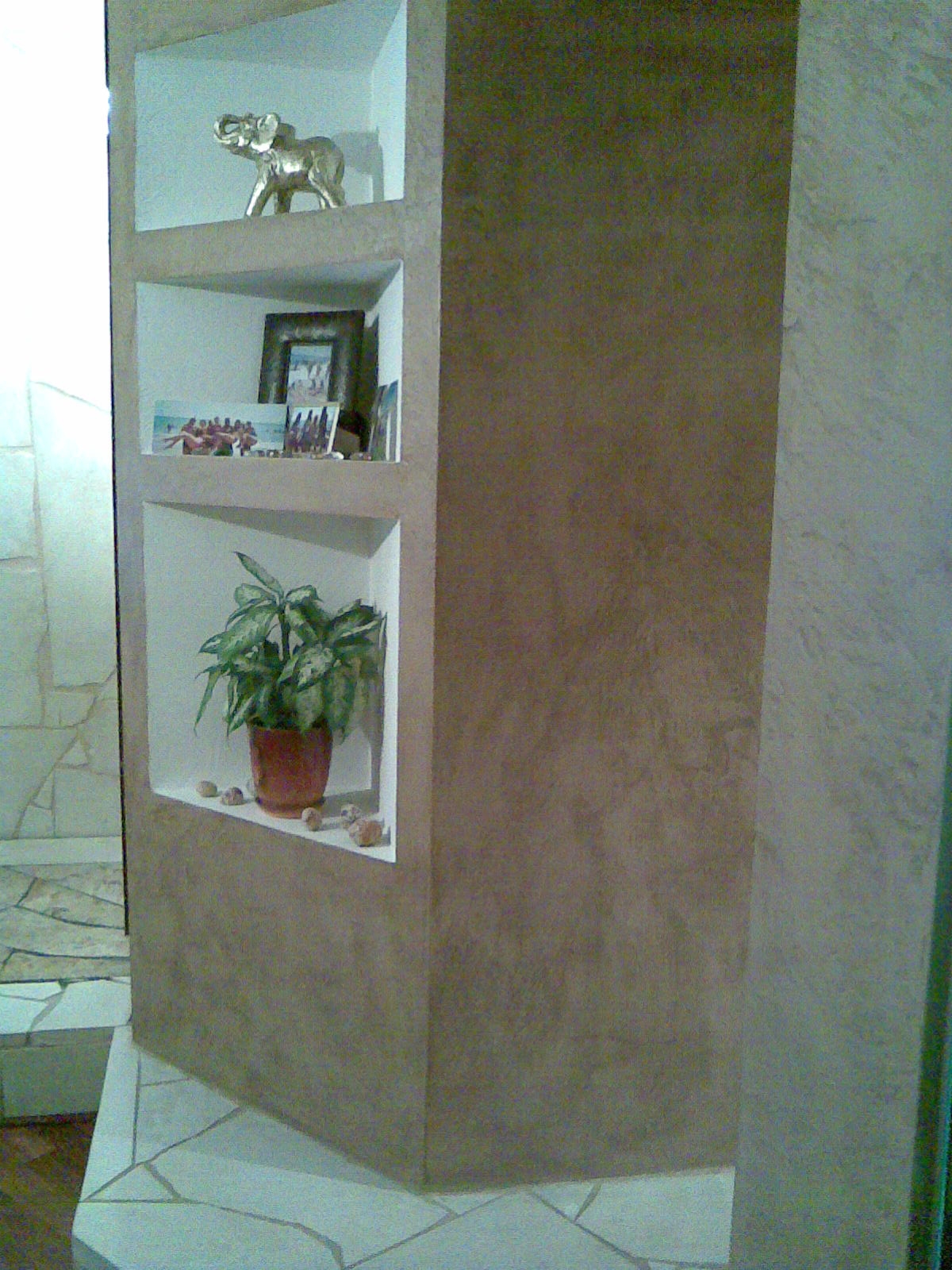 Narożna ściana działowa z trzema wnękami: na górnej złoty słonik, na środkowej ramki ze zdjęciami, na dolnej doniczka z zieloną rośliną, całość wykończona w beżowym tynku strukturalnym.