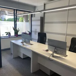 Jasne, nowoczesne biuro z dwoma stanowiskami pracy, białymi biurkami, monitorami Lenovo, czarnymi krzesłami obrotowymi i roślinami w doniczkach przy oknie. Widoczne logo IDEA na biurku.