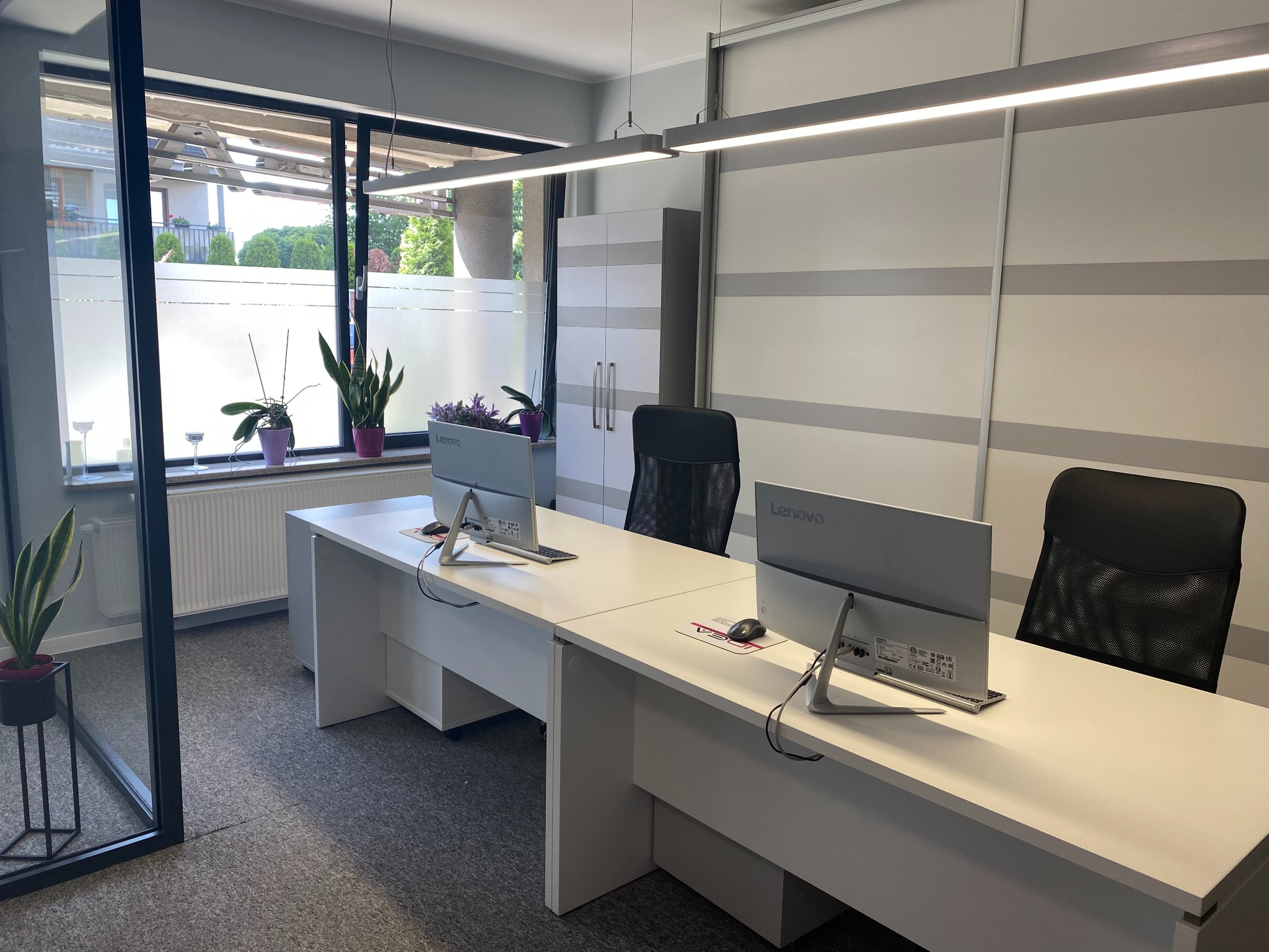 Jasne, nowoczesne biuro z dwoma stanowiskami pracy, białymi biurkami, monitorami Lenovo, czarnymi krzesłami obrotowymi i roślinami w doniczkach przy oknie. Widoczne logo IDEA na biurku.