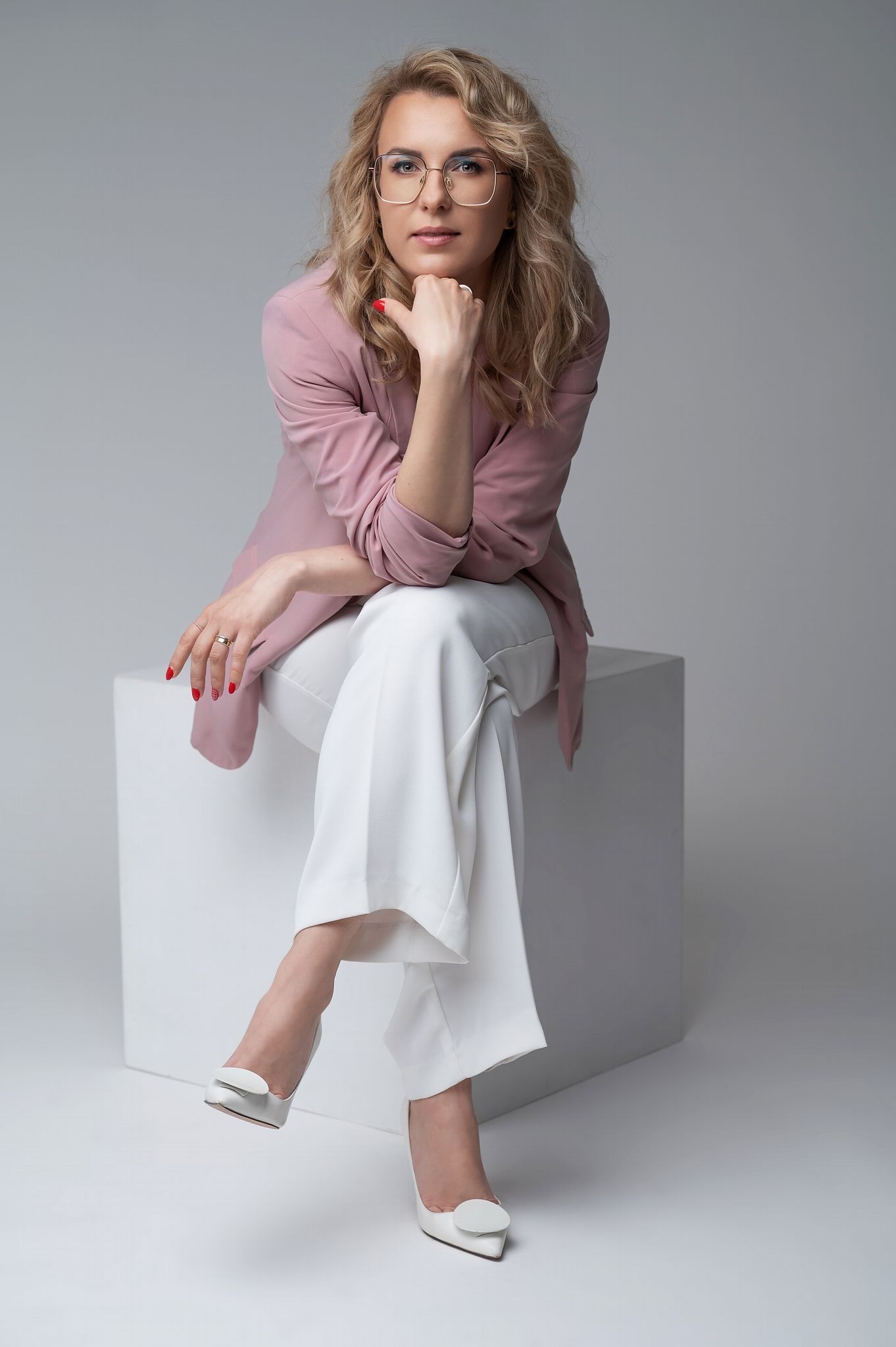 Portret kobiety w okularach siedzącej na białym sześcianie, ubrana w różowy żakiet i białe spodnie, z dłonią podpartą o brodę, na jasnoszarym tle.