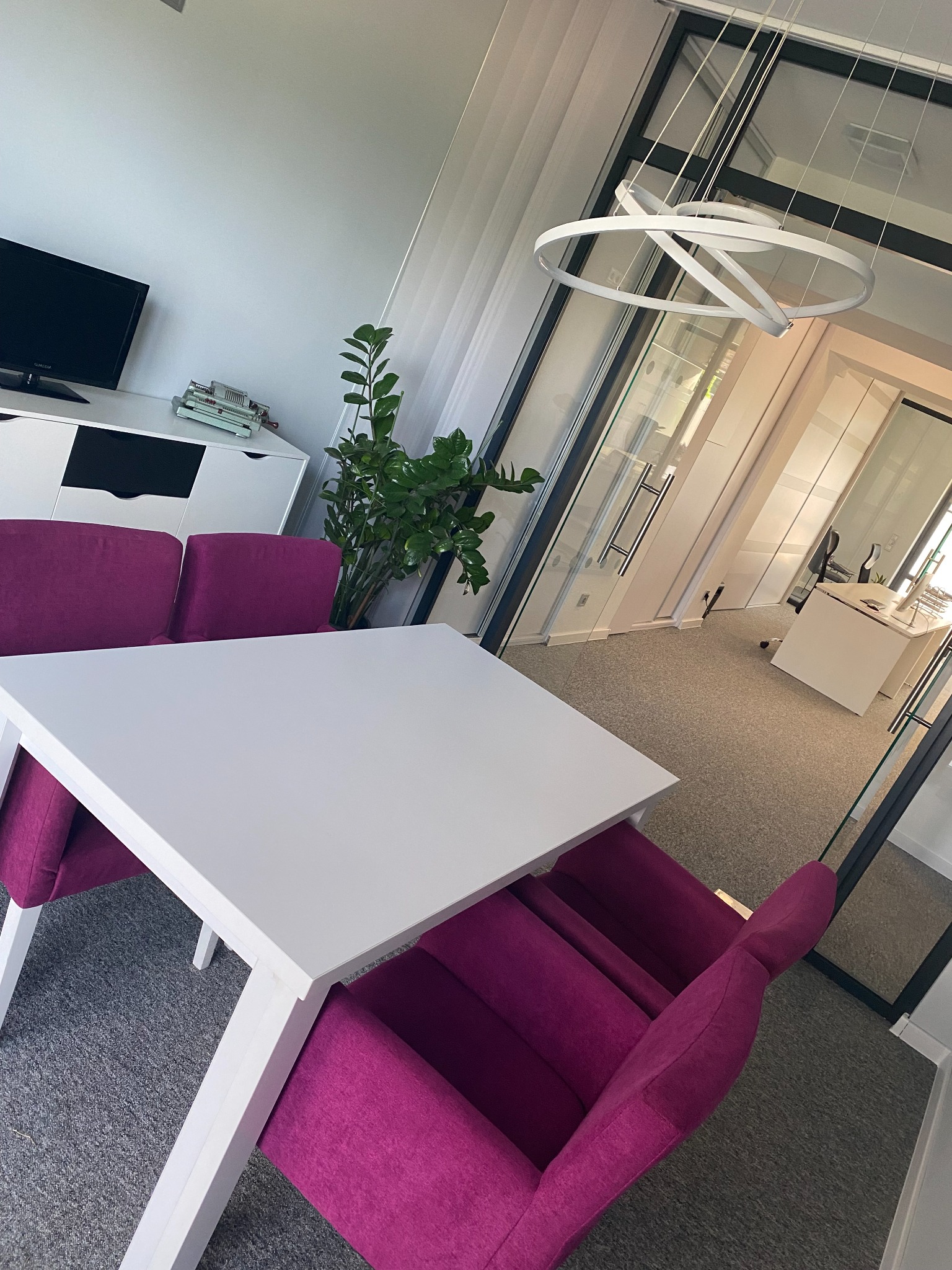 Jasne biuro z białym stołem, fioletowymi krzesłami i sofą, widok na przeszklone biura w tle, nowoczesna lampa sufitowa.