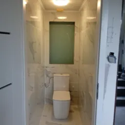 Wąska łazienka w trakcie wykańczania, z toaletą i marmurowymi płytkami na ścianach i podłodze, widoczny niezamaskowany otwór wentylacyjny i niedokończone wykończenie ościeżnicy.