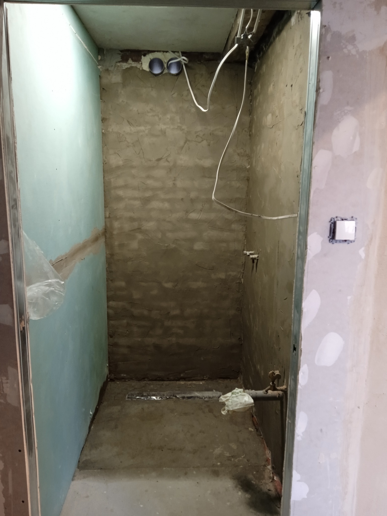 Surowy stan łazienki przed wykończeniem: ściany pokryte szarym tynkiem, widoczne rury i przewody elektryczne, metalowe profile przy ścianach, na podłodze odpływ liniowy.