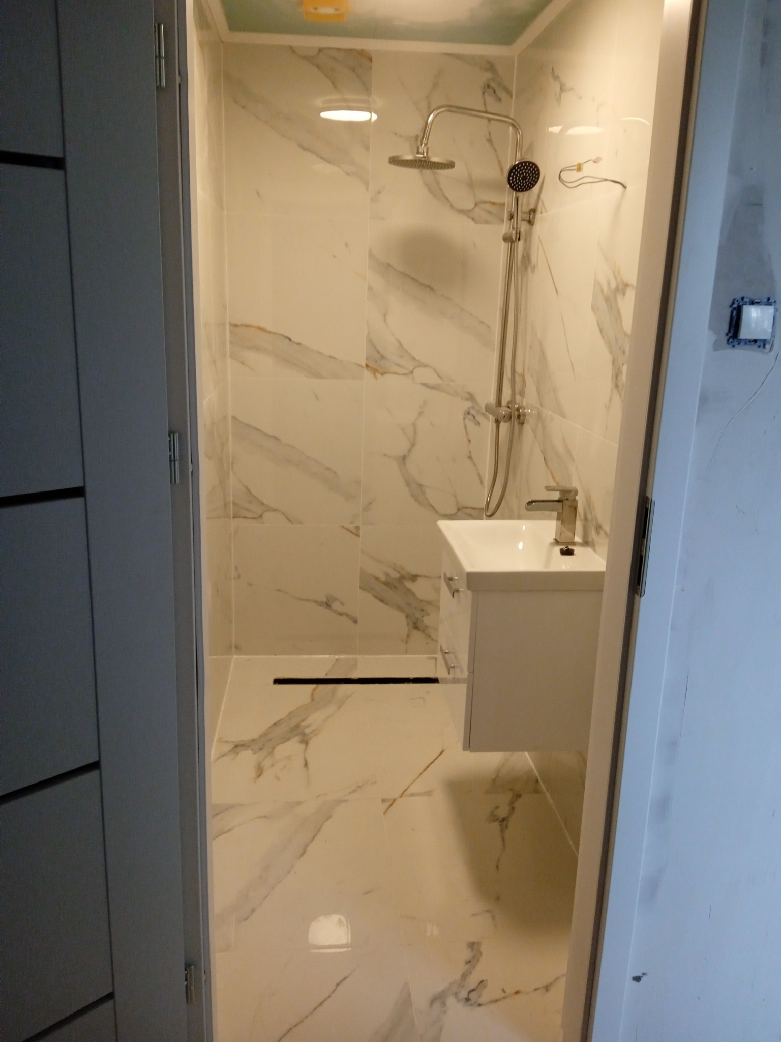 Nowoczesna łazienka z marmurowymi płytkami, kabiną prysznicową typu walk-in i białą szafką umywalkową, widok zza uchylonych drzwi.