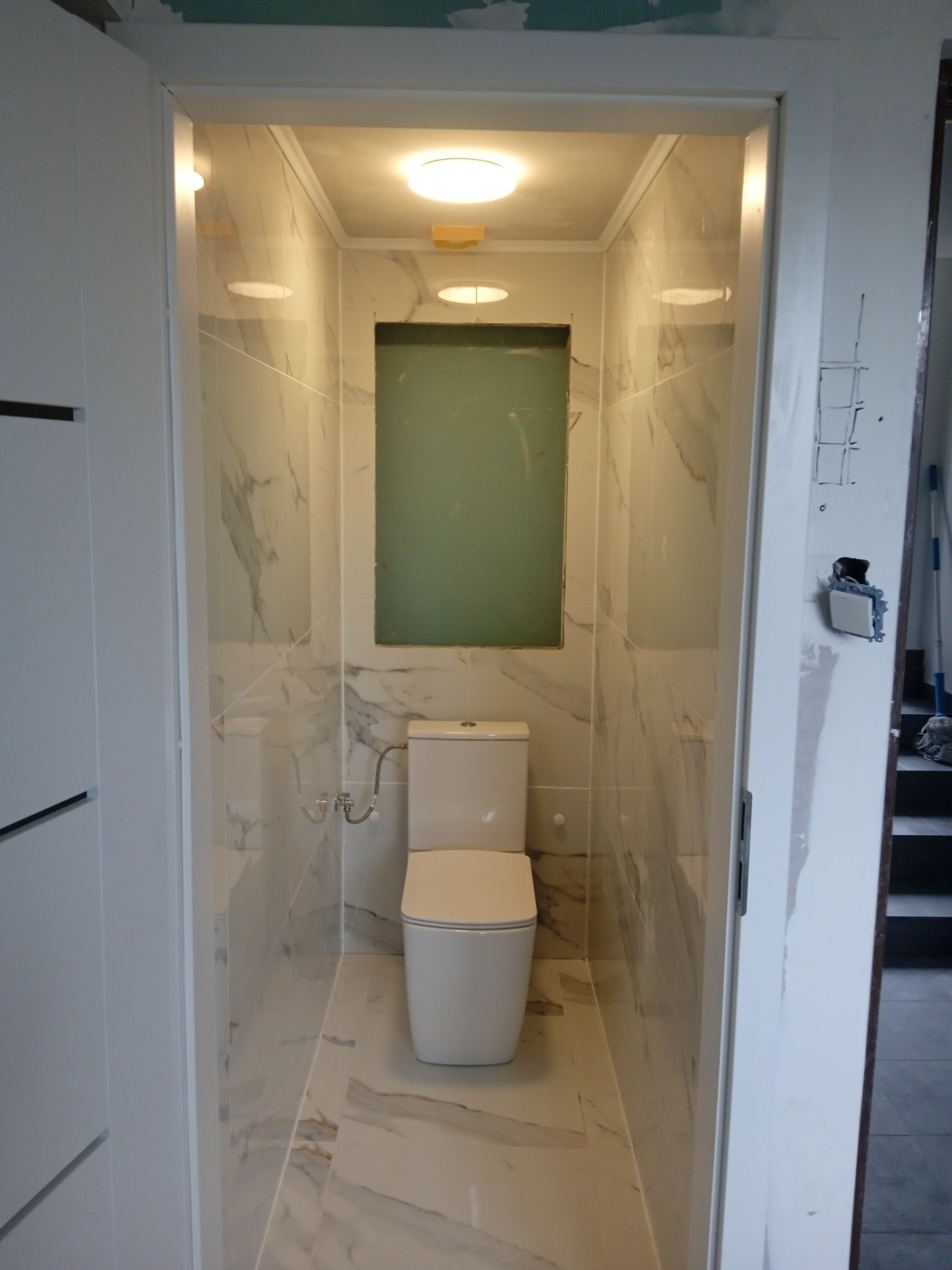 Wąska łazienka w trakcie wykańczania, z toaletą i marmurowymi płytkami na ścianach i podłodze, widoczny niezamaskowany otwór wentylacyjny i niedokończone wykończenie ościeżnicy.