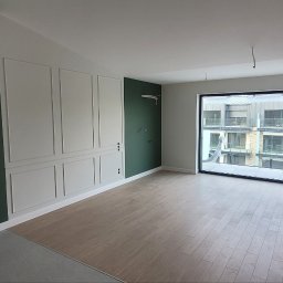 Fedir Melnyk - Wnętrze mieszkania po remoncie, z panelami podłogowymi, ścianami w kolorach szarym i butelkowej zieleni, dekoracyjnymi listwami ściennymi i dużym oknem z widokiem na budynek mieszkalny.