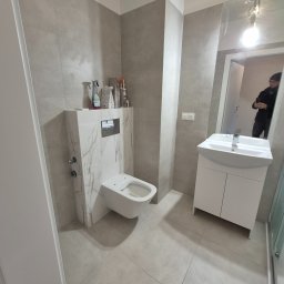 Fedir Melnyk - Nowoczesna łazienka w szarościach z podwieszaną toaletą obudowaną płytkami imitującymi marmur, białą szafką z umywalką i dużym lustrem. Widoczne akcesoria łazienkowe.