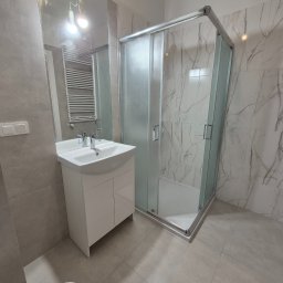 Fedir Melnyk - Jasna łazienka po remoncie: kabina prysznicowa z mleczną szybą, umywalka z szafką, płytki imitujące marmur na ścianach, szare płytki podłogowe i wiszące żarówki.