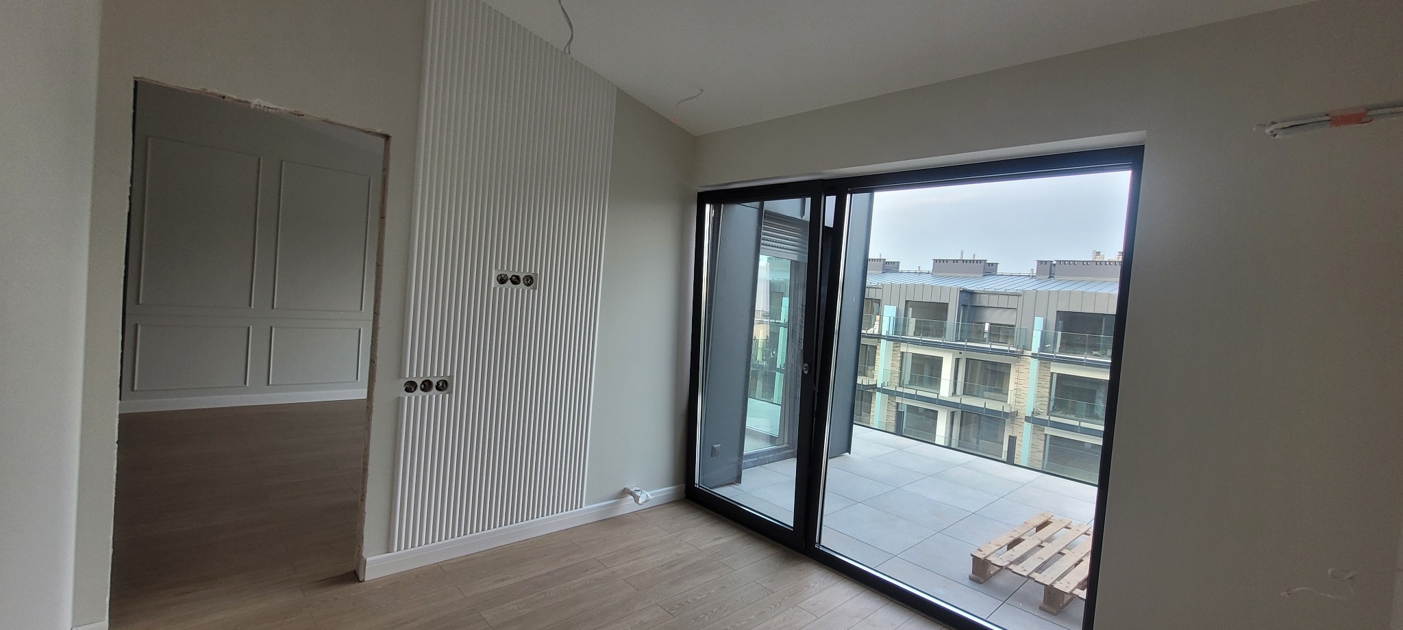 Wnętrze mieszkania w trakcie wykańczania: ściana z dekoracyjnymi panelami pionowymi i gniazdkami elektrycznymi, widok na balkon z paletą i budynki mieszkalne przez duże okno przesuwne, w tle...