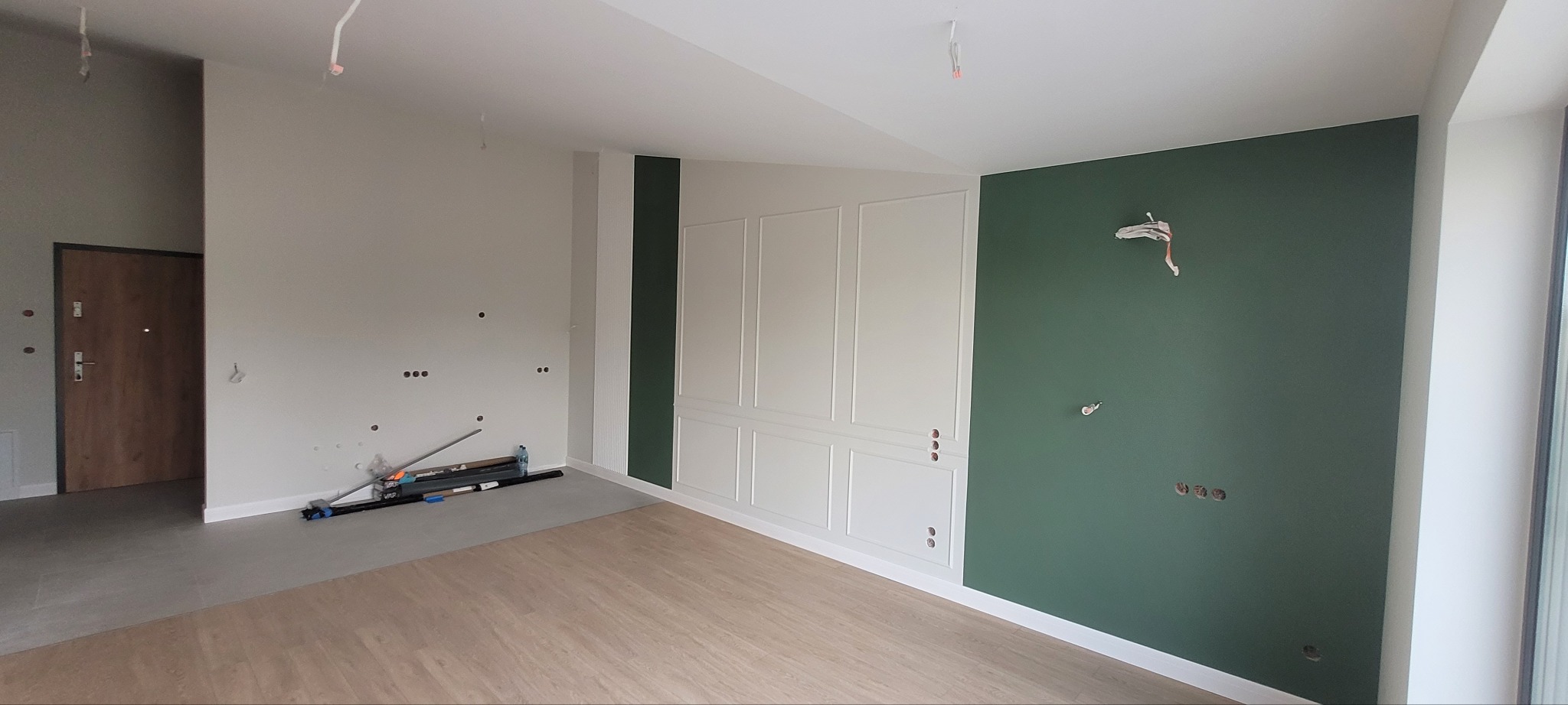 Pokój w trakcie remontu: ściana pomalowana na zielono, obok biała ściana z dekoracyjnymi panelami, widoczne puszki elektryczne, podłoga częściowo wyłożona panelami, narzędzia na podłodze.