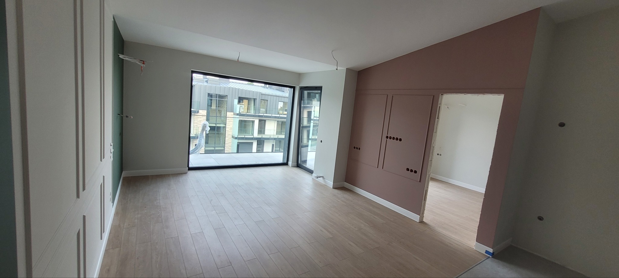 Wnętrze pokoju po remoncie: świeżo pomalowane ściany w odcieniach beżu i zieleni, ułożone panele podłogowe, duże okno z widokiem na budynek mieszkalny. Widoczne niedokończone prace elektryczne.