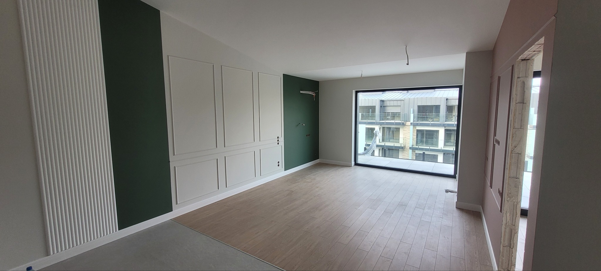 Wnętrze mieszkania po remoncie, z panelami podłogowymi, ścianami w kolorach szarym i butelkowej zieleni, dekoracyjnymi listwami ściennymi i dużym oknem z widokiem na budynek mieszkalny.