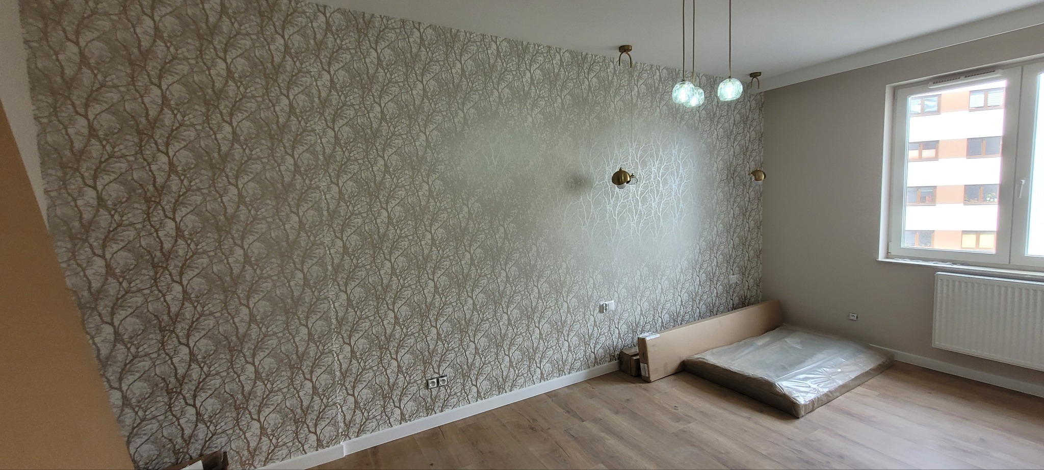 Pokój po remoncie: ściana pokryta tapetą w motywy drzew, drewnopodobna podłoga, nowoczesne oświetlenie sufitowe i ścienne, okno z widokiem na budynek mieszkalny, grzejnik, kartonowe opakowania...