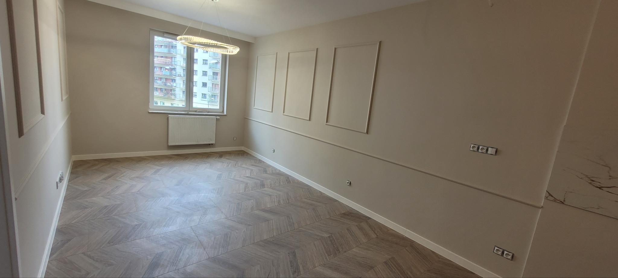 Jasne wnętrze pokoju po remoncie, z parkietem ułożonym we wzór jodełki, ozdobnymi panelami na ścianach i nowoczesną lampą wiszącą.