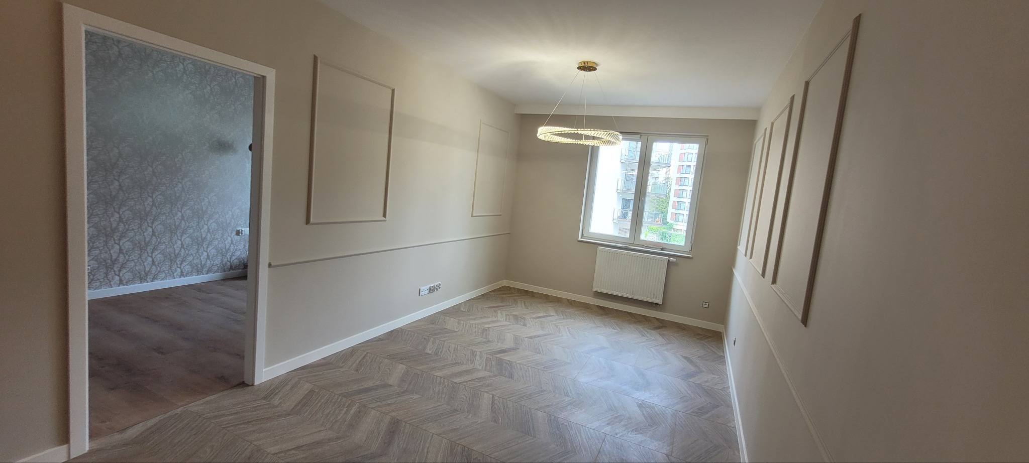 Jasne wnętrze po remoncie: parkiet w jodełkę, beżowe ściany z dekoracyjnymi listwami, okno z widokiem na budynek, nowoczesna lampa wisząca