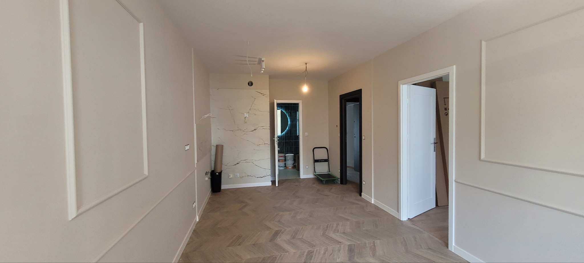 Wykończone wnętrze mieszkania z parkietem w jodełkę, kremowymi ścianami z dekoracyjnymi listwami, widoczne drzwi w kolorze białym i czarnym, oraz fragment łazienki z podświetlanym lustrem