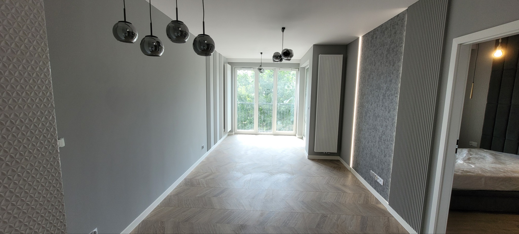 Nowoczesny salon z szarymi ścianami, parkietem ułożonym w jodełkę, oświetleniem kulistym i dekoracyjną ścianą z pionowymi panelami, widok na balkon i sypialnię.