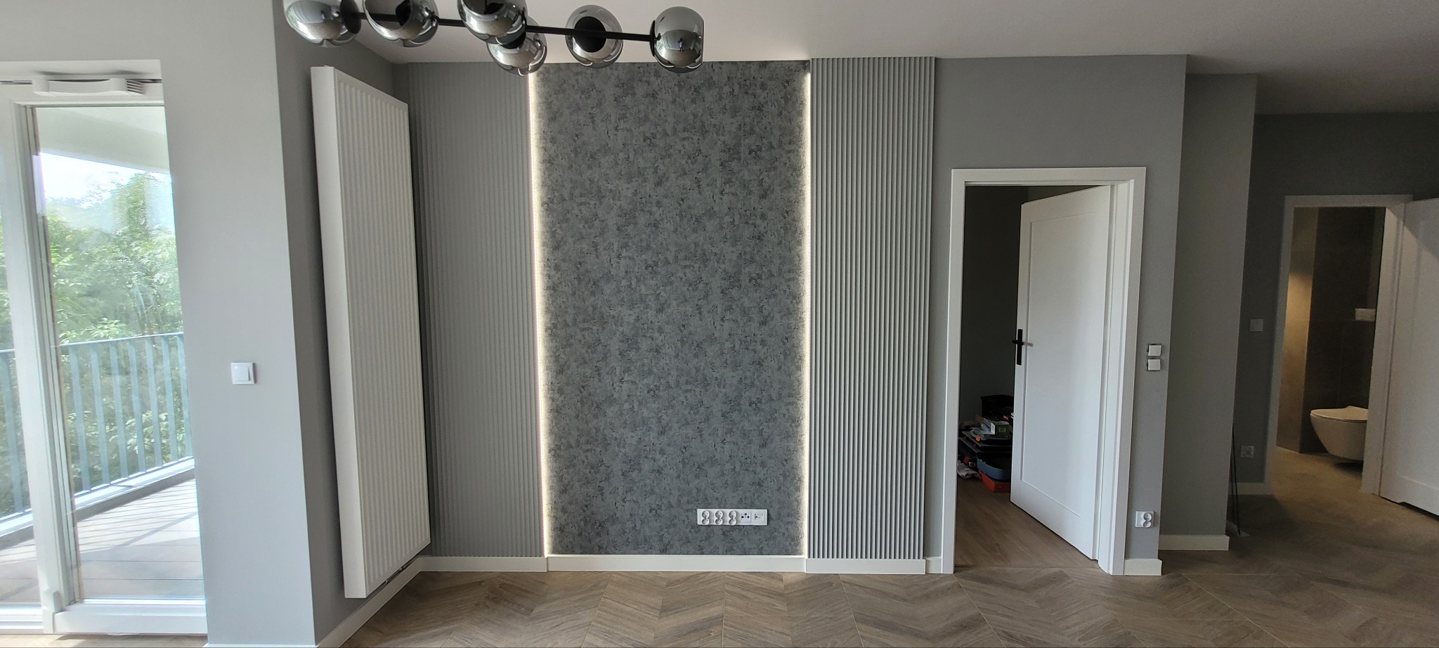 Nowoczesne wnętrze mieszkania z panelami dekoracyjnymi na ścianie, oświetleniem LED, grzejnikiem panelowym oraz podłogą w jodełkę, widok na balkon i otwarte drzwi do pomieszczeń.