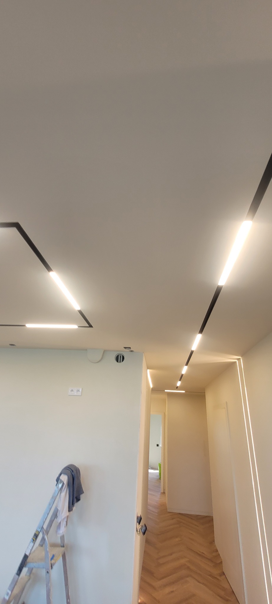 Nowoczesne oświetlenie sufitowe w trakcie montażu, widoczna drabina malarska i fragment podłogi z parkietem w jodełkę.