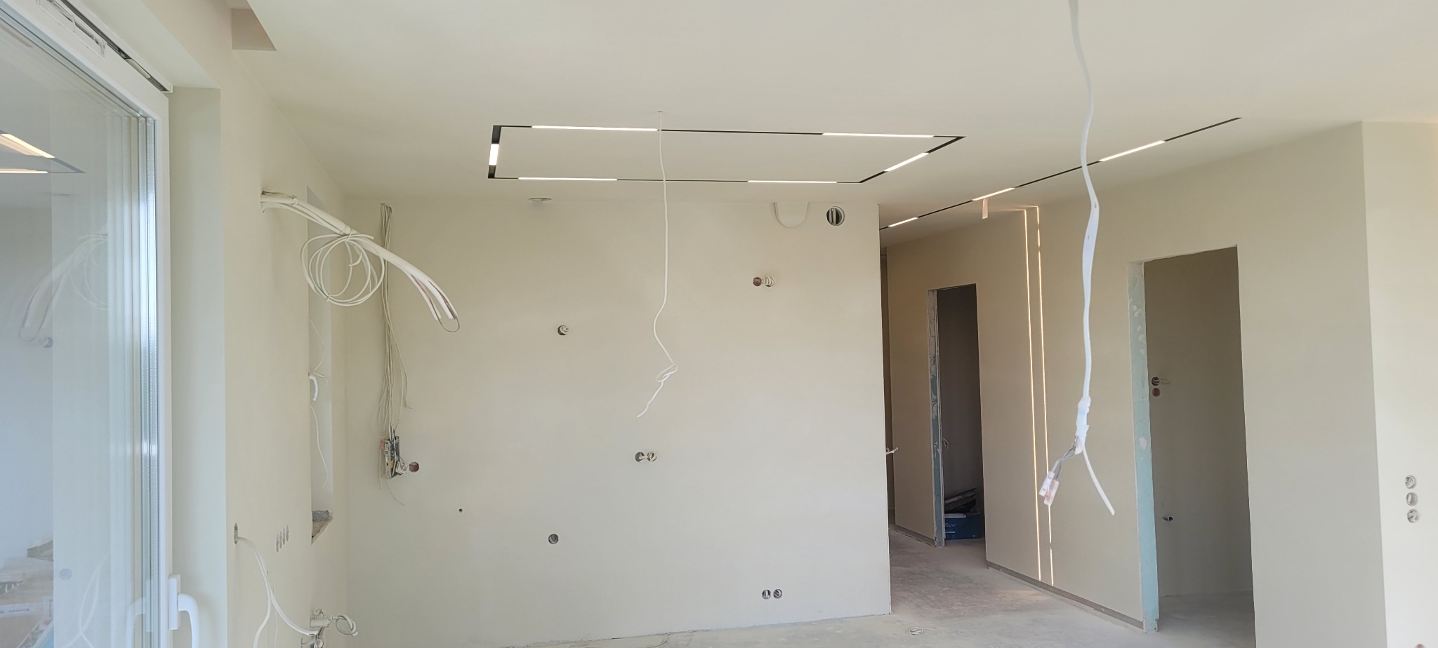 Wnętrze w trakcie prac wykończeniowych; widoczne przewody elektryczne zwisające ze ściany i sufitu, zamontowane oświetlenie LED w suficie i ścianach, surowe ściany przygotowane do malowania, otwory...