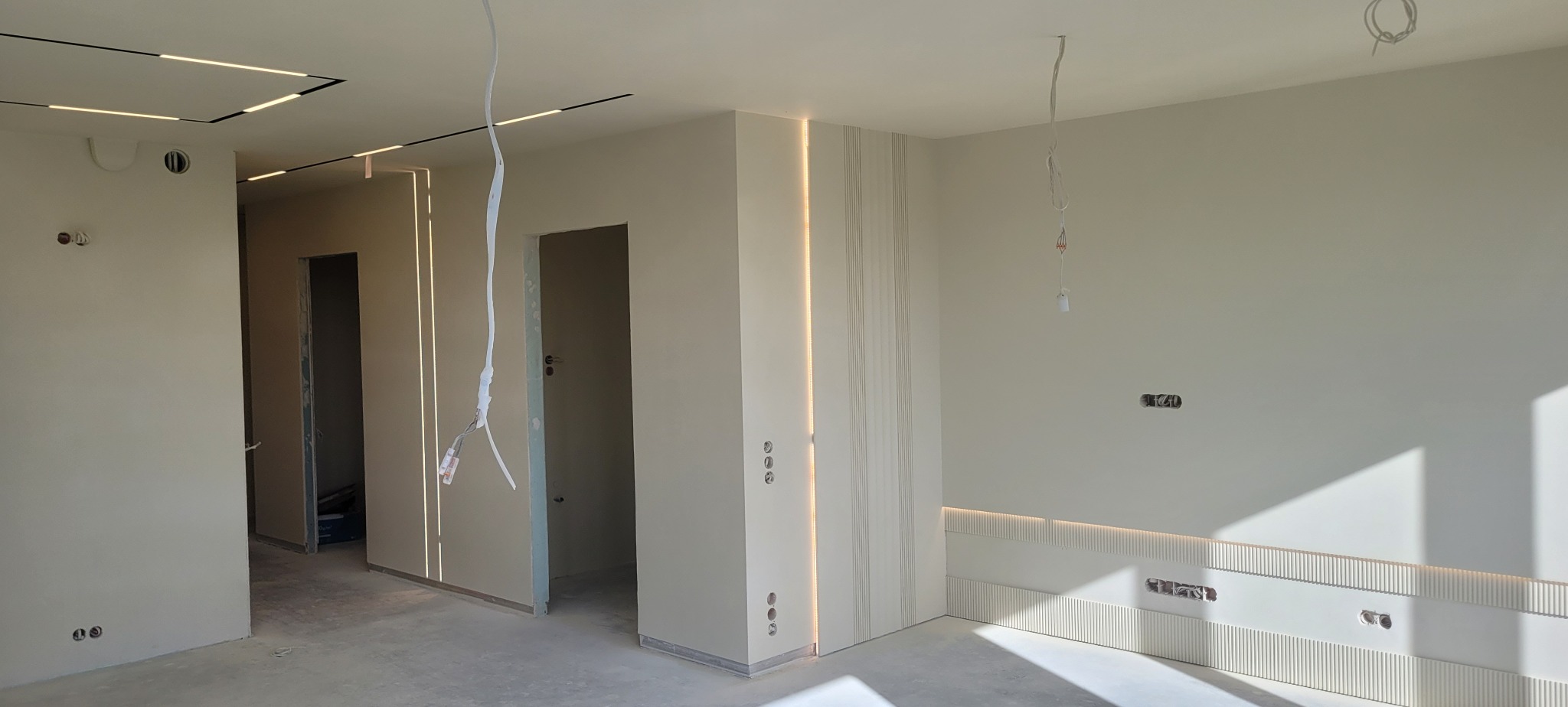 Jasne wnętrze w trakcie prac wykończeniowych, widoczne przygotowane ściany, oświetlenie LED w suficie i pionowe panele dekoracyjne z podświetleniem, niezabezpieczone przewody elektryczne zwisają...