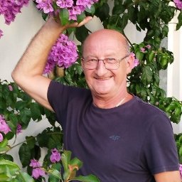 DZIADEK TADEK TADEUSZ WĄS - Uśmiechnięty mężczyzna w okularach pozuje przy kwitnącej bugenwilli, trzymając gałązkę z kwiatami w dłoni.